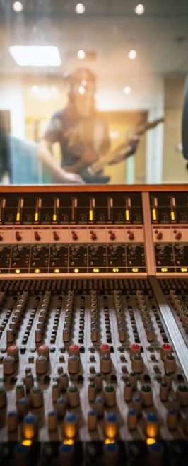 A close-up of a professional mixing desk with glowing indicator lights, with a blurred guitarist visible through the studio window A close-up of a professional mixing desk with glowing indicator lights, with a blurred guitarist visible through the studio window